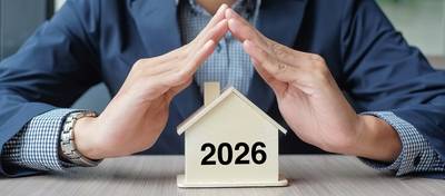 Geschäftsmann übergibt hölzernes Hausmodell mit Neujahrstext 2026. Immobilienversicherung und Immobilienkonzepte