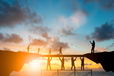 Menschen laufen über eine Brücke, die von anderen Personen von unten abgestützt wird in eindrucksvoller Natur mit Wolkenhimmel und Sonnenstrahlen als Zeichen für Teamwork.