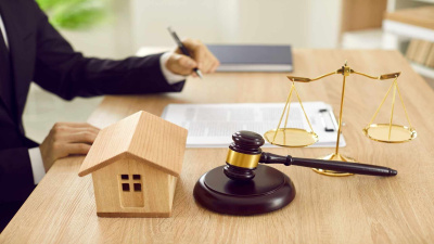 Anwalt sitzend am Schreibtisch mit Waage der Justiz, kleinem Holzspielhaus und Arbeitsdokumenten drauf.terzeichnung von Verträgen. 