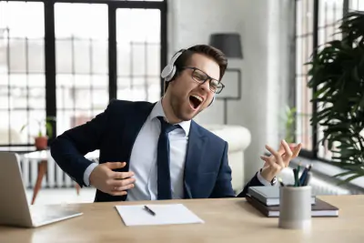 Business-Mann mit Kopfhörern, der so tut, als spiele er Gitarre, sitzt am Schreibtisch im modernen Büro