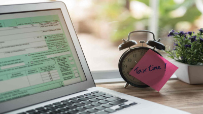 Notebook mit Steuerformular auf dem Bildschirm, daneben ein Wecker mit Post-it auf dem Tax Time steht.