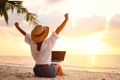 Rückansicht auf junge Frau mit Strohhut, die auf einem Laptop arbeitet und die Arme hochhält mit Blick auf einen Sonnenuntergang an einem tropischen Sandstrand.