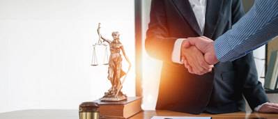 Symbolfoto Vertrag: Zwei Geschäftsleute geben sich die Hand, Blick auf Schreibtisch mit Richterhammer, Dokument und einer Justitia-Figur die auf einem Buch steht