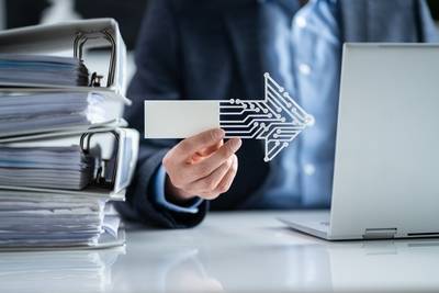 Aktenordner und ein Laptop, dazwischen hält eine Hand einen Pfeil, der die Digitalisierung der Akten symbolisieren soll